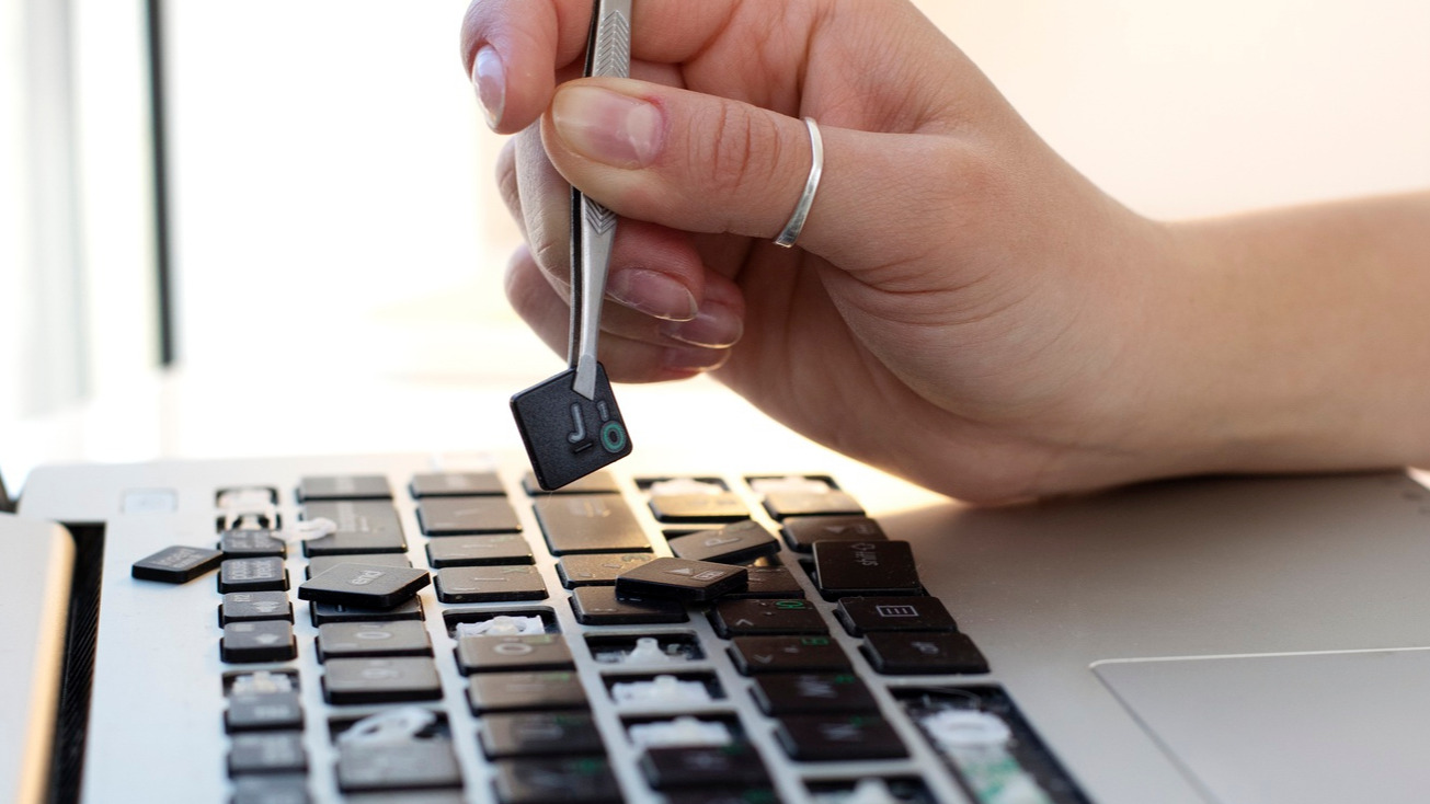 A mão de uma mulher segurando uma tecla com pinças acima de um teclado de laptop com várias teclas faltando.