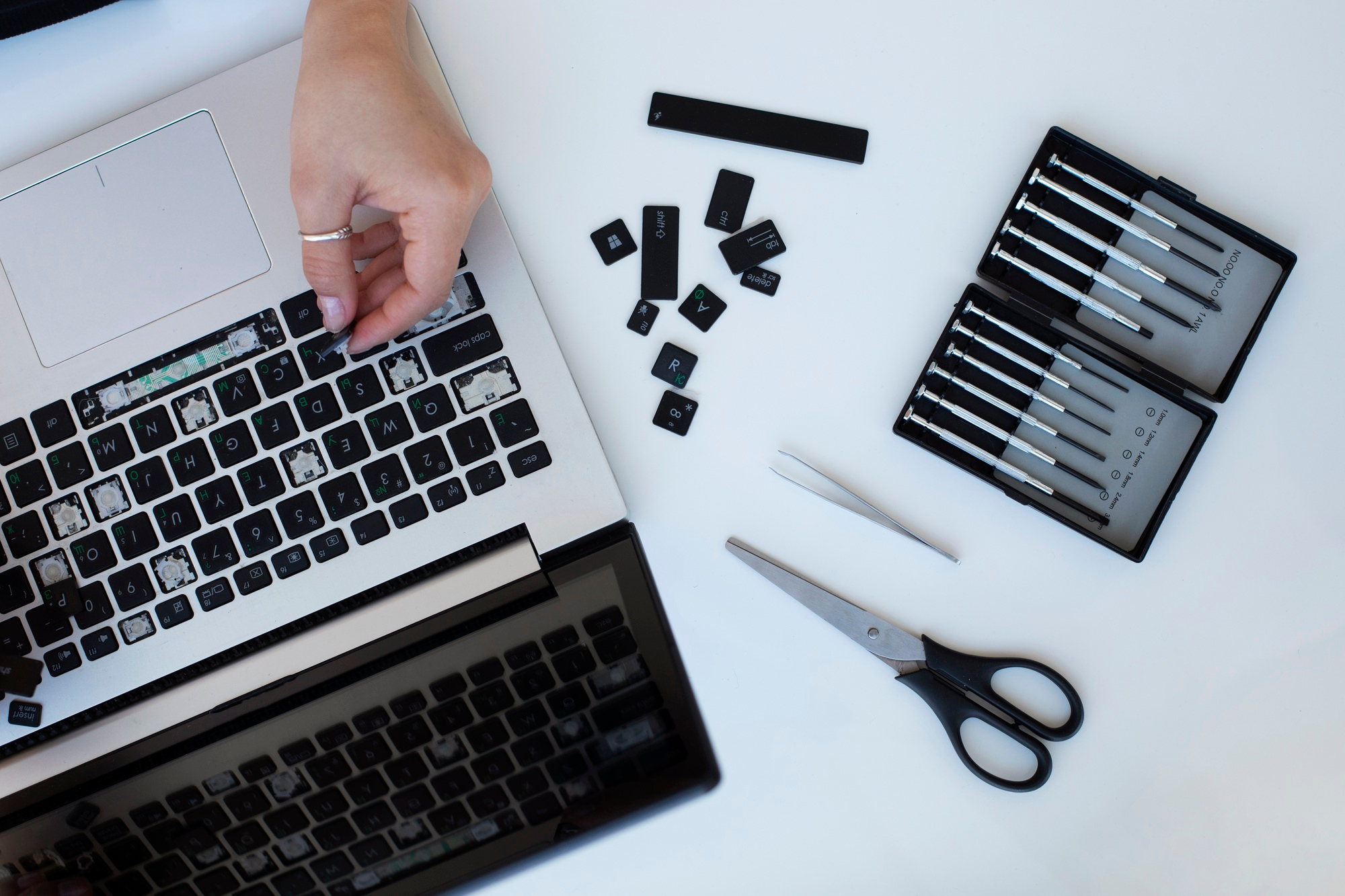 Uma teclado de laptop com teclas removidas, ferramentas e a mão de uma pessoa trabalhando em seu conserto.